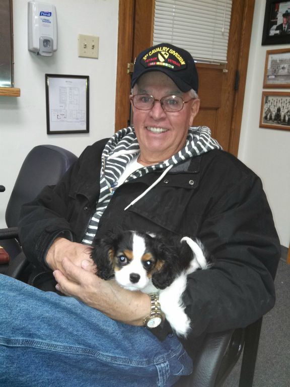 Smiling man in black jacket and glasses, with 1st Cavalry Division hat on, holds small spaniel in lap